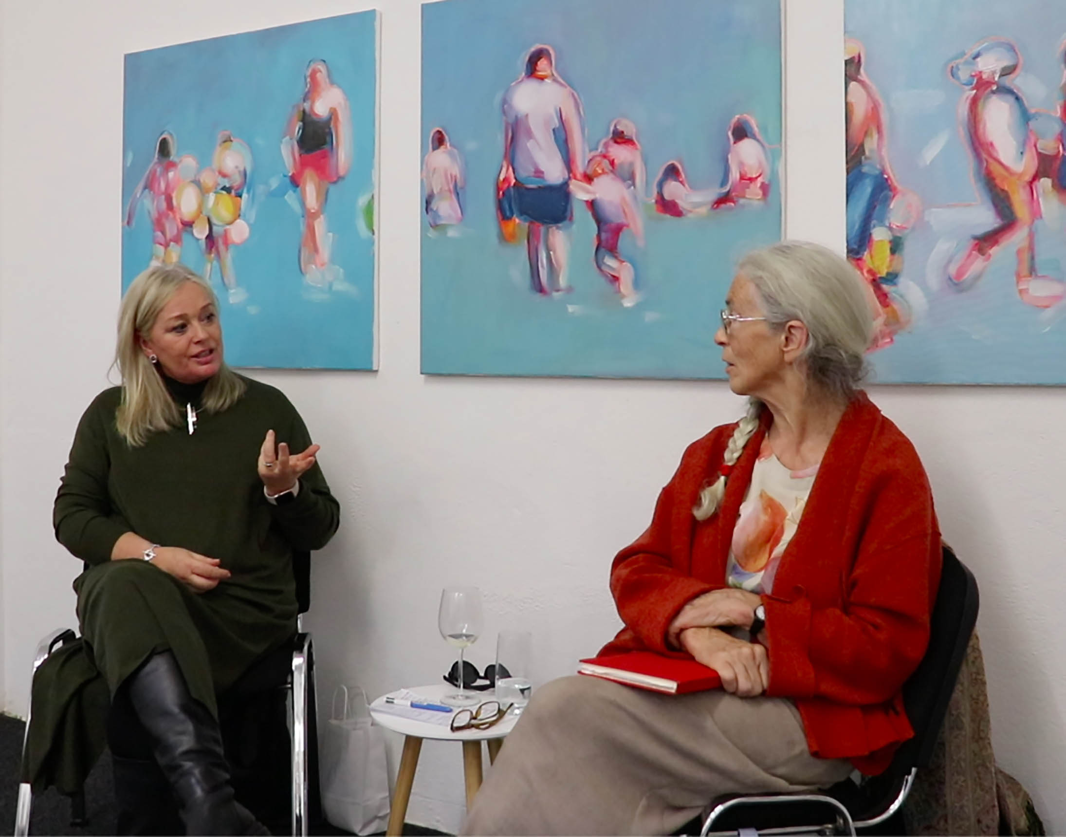 A photo of artist Oonagh Latchford in conversation with art historian and curator Catherine Marshall, in front of three of Oonagh's paintings. The artworks are of abstracted beach scenes, people playing, walking and just standing taking in the sea.